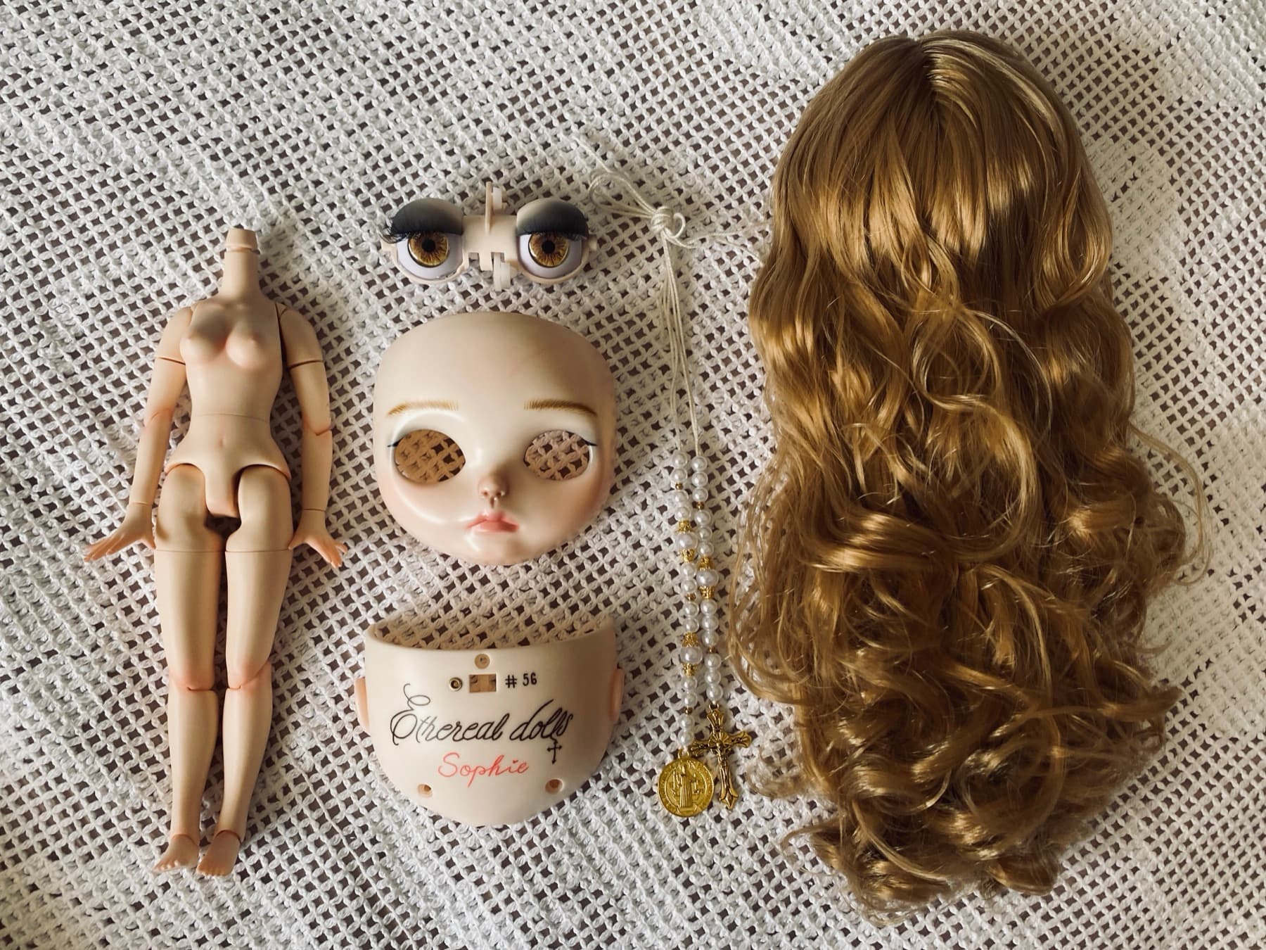 Doll parts laid out: body, painted face plate, eye mechanism, rosary pullstring and wig